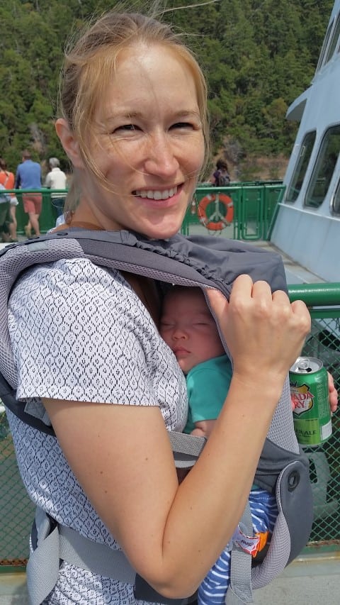 mom and infant on a ferry in Washington State.  Baby is sleeping in the carrier, and mom is smiling. 