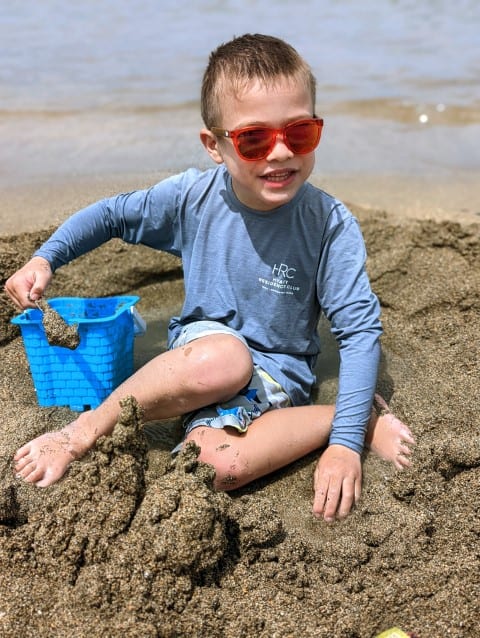 Child wearing orange Knockaround sunglasses and sitting in the sand smiles at the camera