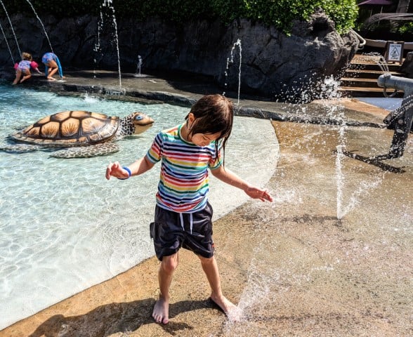 Child wearing a rainbow swim top and grey shorts is playing on a splash pad
