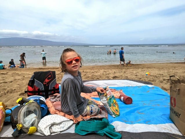 A child is sitting on a beach mat, surrounded by beach gear, is eating chips.  In the background, Maui's Kapalua Beach is visible.