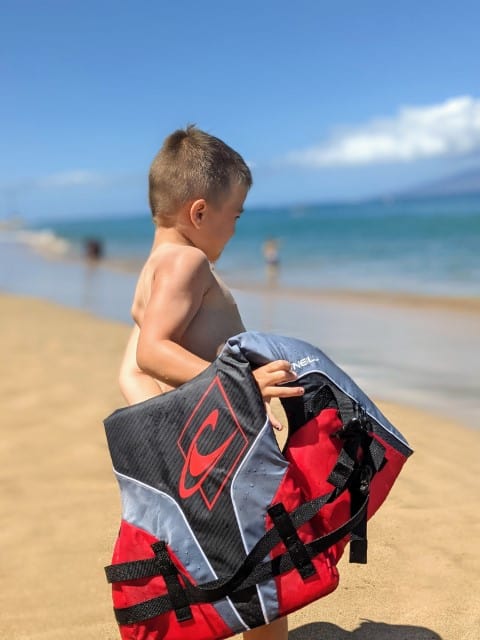 A child is slipping on a red life jacket.  In the background, the blue water of Kaanapali Beach in Maui is visible. 