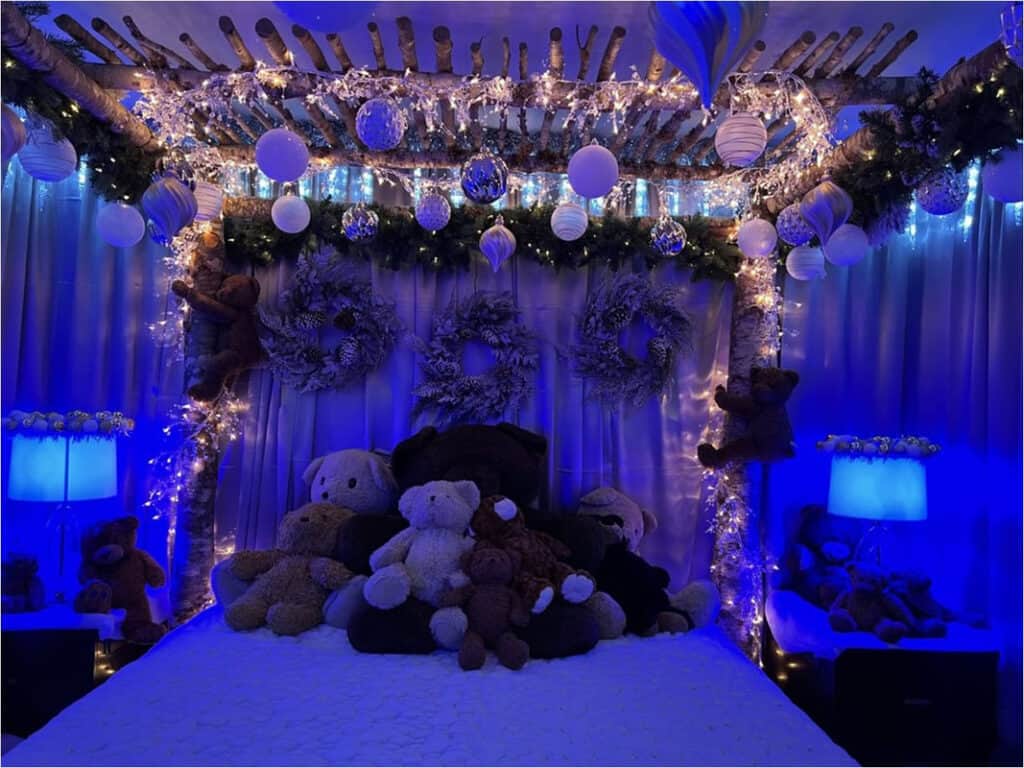 A room with blue lighting. There is a bed with tons of teddy bears sitting at the top. The bed frame is decorated in lights and colorful bulbs. 
