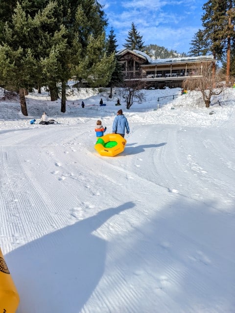 A child and parent walk up a snow covered hill dragging a large yellow and green tube. 