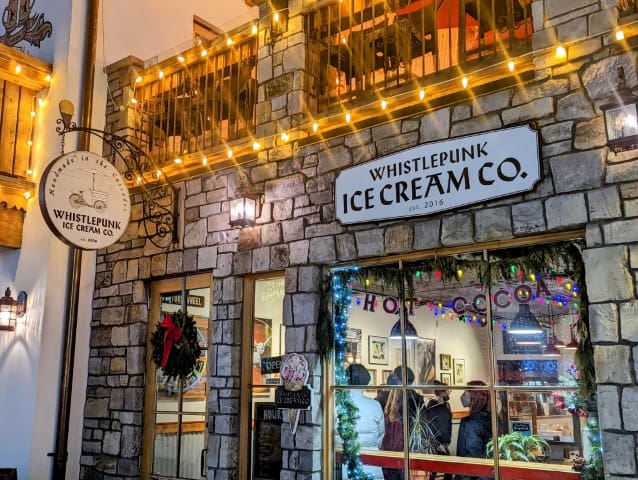 View of the front of the Whistlepunk Ice cream Co shop.  The window is linked with garland and has a sign that reads "hot cocoa".  The outside of the building is grey stone bricks with a railing from an balcony above. 