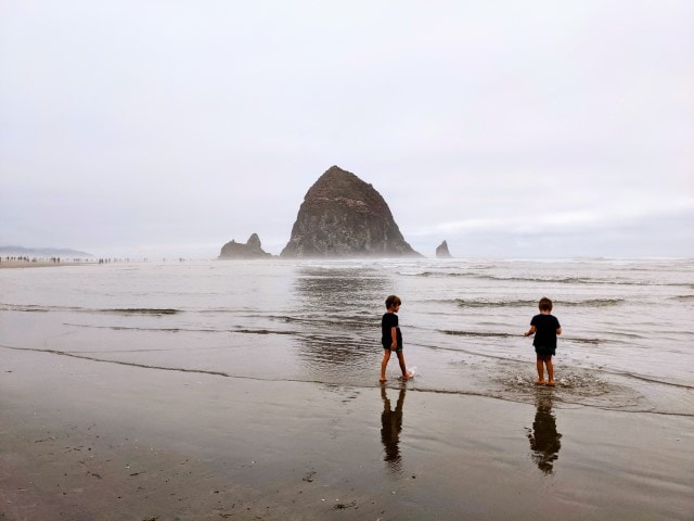 Two kids walking in the water in front of  the famous Canon Beach Haystack